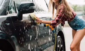 Uśmiechnięta kobieta myje auto gąbką i wężem. Domowe sposoby na czysty samochód to świetna zabawa!