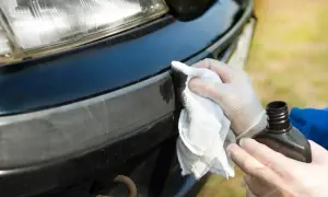 Ręce w rękawiczkach aplikują środek do szlifowania lakieru na zderzak samochodu, przygotowując go do dalszych prac.