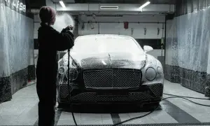 Osoba w czarnym kombinezonie spryskuje luksusowe auto pianą podczas profesjonalnego detalingu auta w myjni.