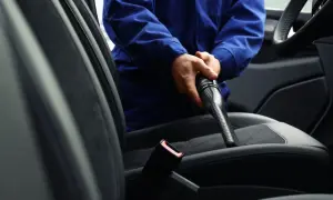 Odkurzanie wnętrza samochodu. Pracownik w niebieskim kombinezonie używa odkurzacza do czyszczenia siedzeń.