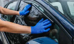 Ręce w niebieskich rękawiczkach usuwają pogniecioną folię z ciemnego okna samochodu. Poradnik: jak usunąć folię z okna.