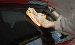 Ręka z pomarańczową mikrofibrą czyści tylną szybę czerwonego auta. W tle widać spryskiwacz, pokazujący, jak myć auto na myjni.