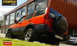 Pomarańczowy SUV z czarnymi dodatkami, jak baranek na tle nowoczesnego budynku. Zapasowe koło z tyłu.