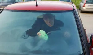 Mężczyzna w samochodzie próbuje pozbyć się wilgoci z zaparowanej szyby, używając zielonej ściereczki.