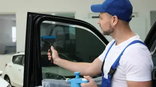 Mężczyzna w niebieskiej czapce i ogrodniczkach przyciemnia szyby samochodu, aby uniknąć mandatu za przyciemnione szyby.