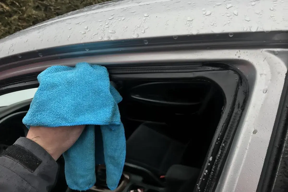 Ręka z niebieską ściereczką czyści uszczelki drzwi samochodu. Dowiedz się, czym smarować uszczelki w samochodzie domowe sposoby, by zapobiec skrzypieniu.