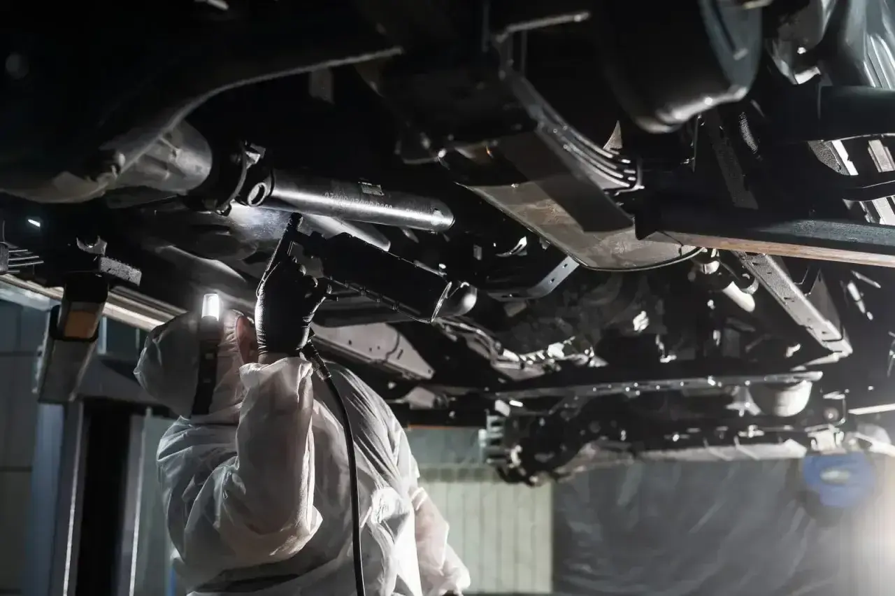 Mechanik w kombinezonie ochronnym wykonuje konserwację podwozia bez kanału, używając pistoletu do aplikacji środka.