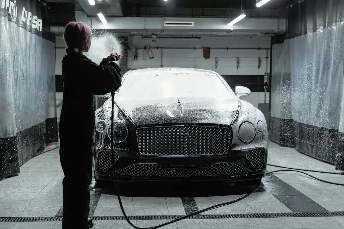 Osoba w czarnym kombinezonie spryskuje luksusowe auto pianą podczas profesjonalnego detalingu auta w myjni.