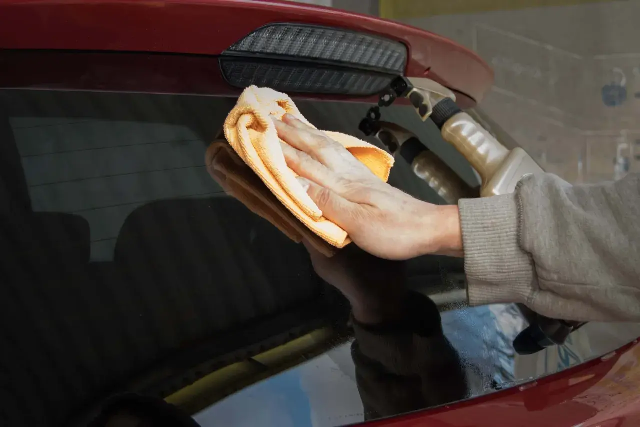 Ręka z pomarańczową mikrofibrą czyści tylną szybę czerwonego auta. W tle widać spryskiwacz, pokazujący, jak myć auto na myjni.