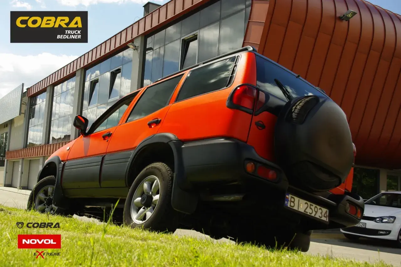 Pomarańczowy SUV z czarnymi dodatkami, jak baranek na tle nowoczesnego budynku. Zapasowe koło z tyłu.