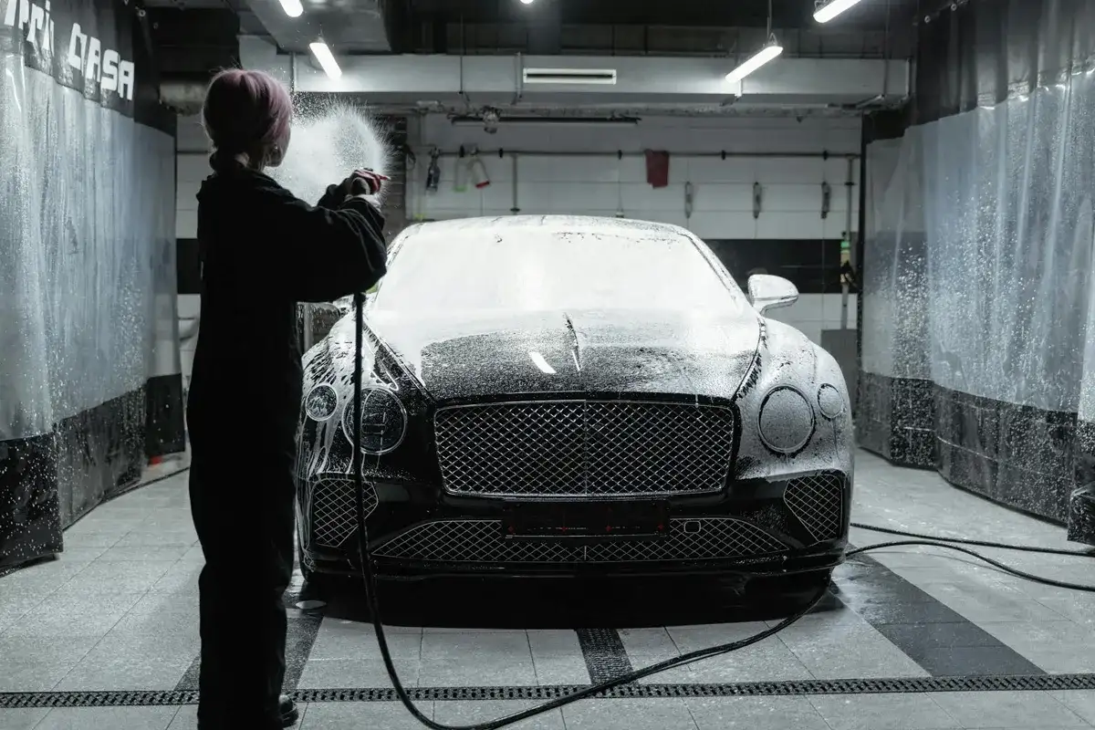 Osoba w czarnym kombinezonie spryskuje luksusowe auto pianą podczas profesjonalnego detalingu auta w myjni.