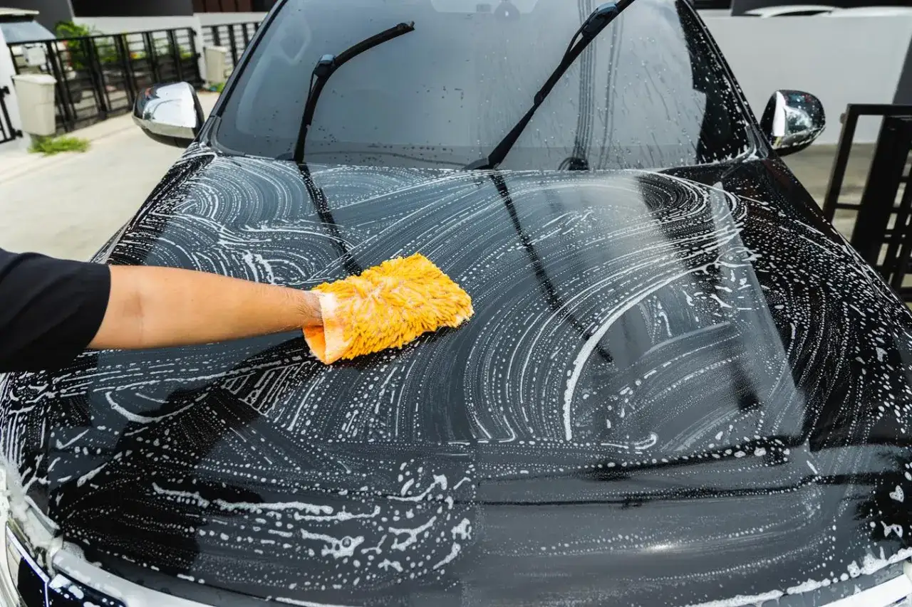 Ręka w pomarańczowej rękawicy do mycia wykonuje mycie detailingowe czarnego samochodu, pokrytego pianą.