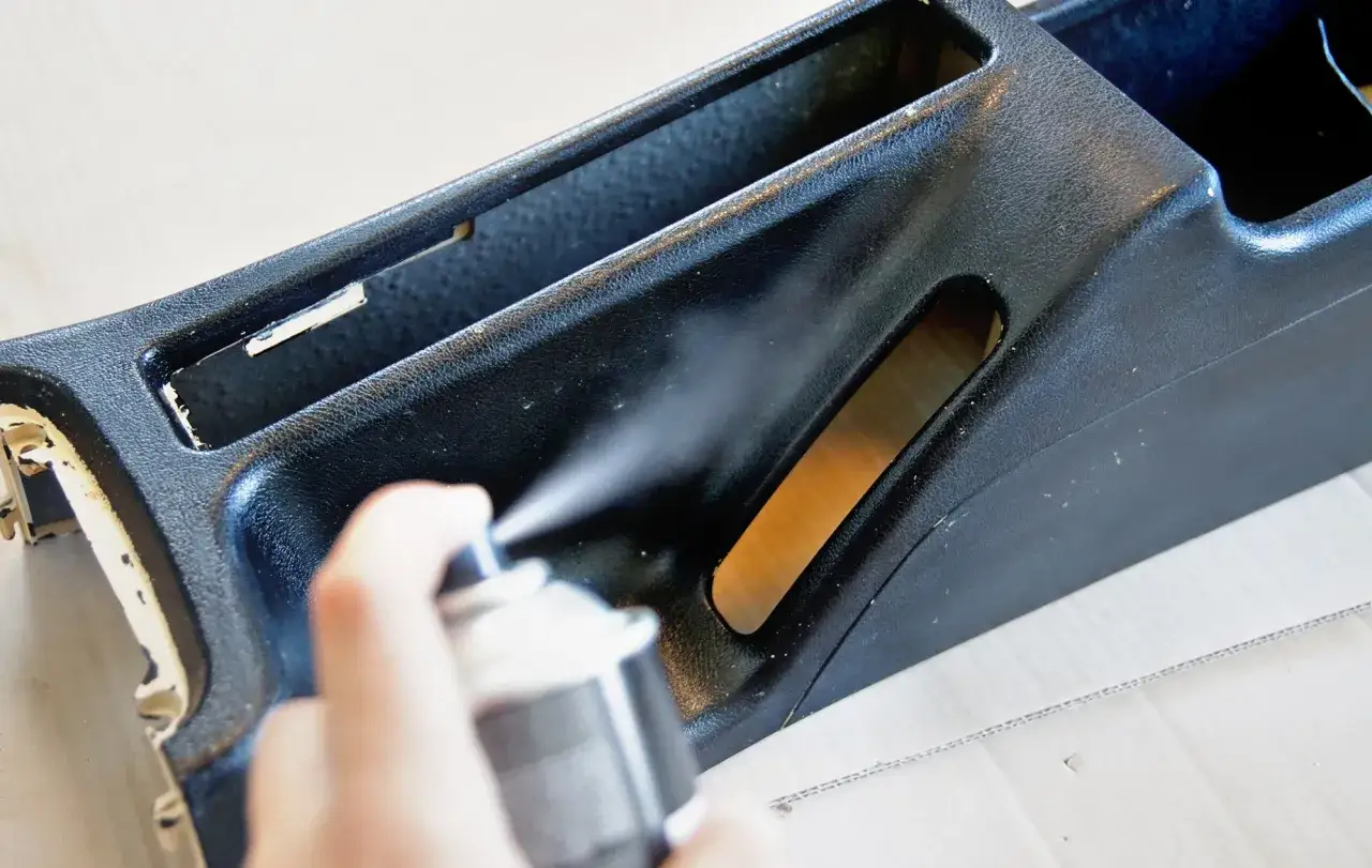 Ręka trzyma puszkę z aerozolem, odświeżając czarny plastikowy element konsoli. Proces regeneracji plastików w toku.