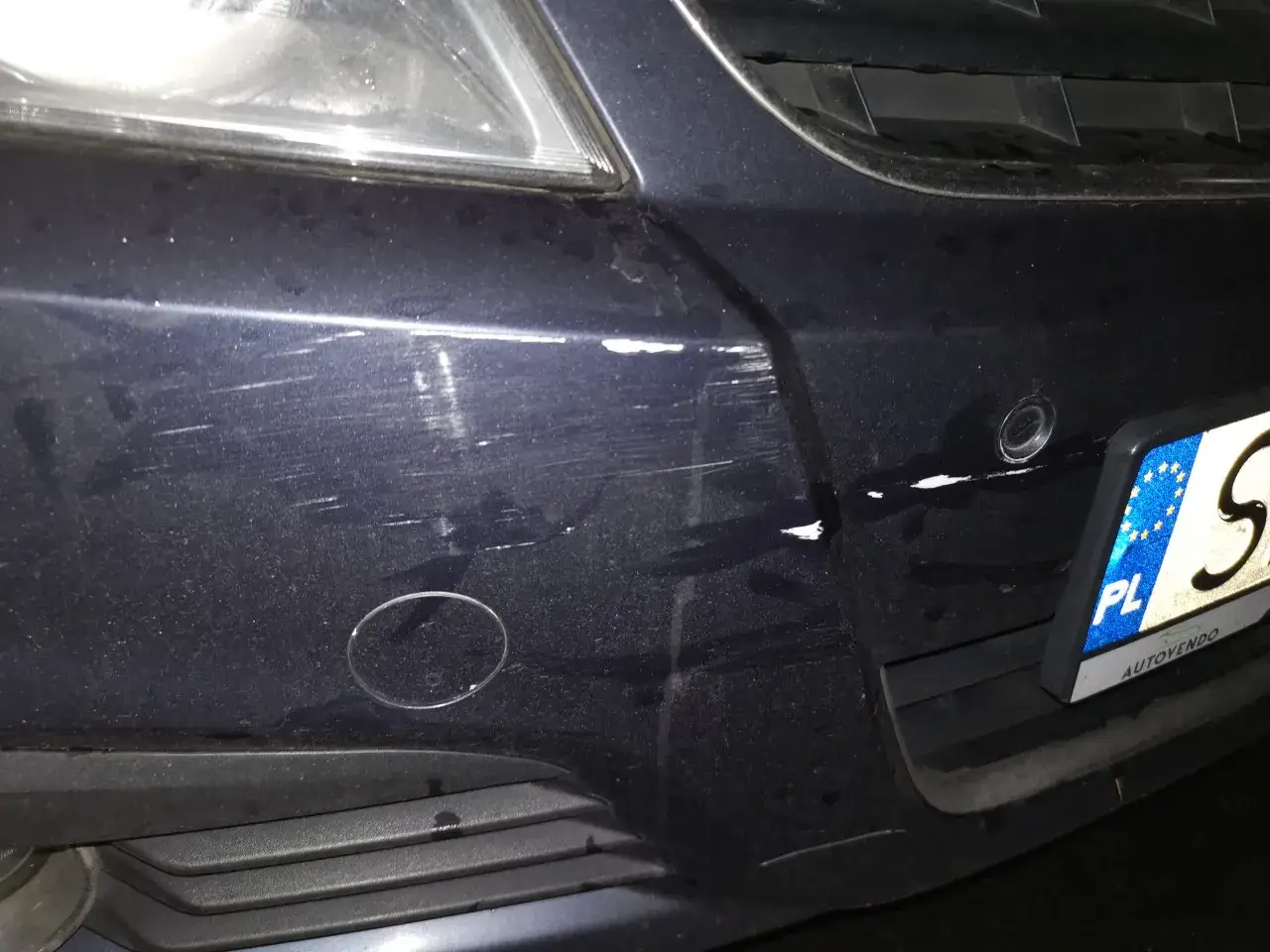 Zderzak z licznymi zadrapaniami i odpryskami, widoczny zdarty lakier. Widać czujniki parkowania i fragment tablicy rejestracyjnej.